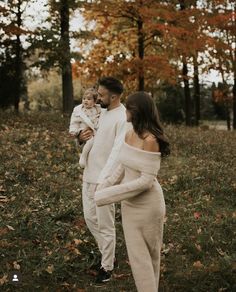 a man and woman holding a baby in their arms while walking through the woods with leaves all around them