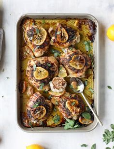 chicken with lemons and herbs in a baking dish on a white table next to some lemons