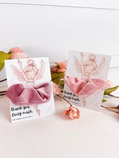 two cards with pink bows on them sitting next to some flowers and leaves in front of a white wall