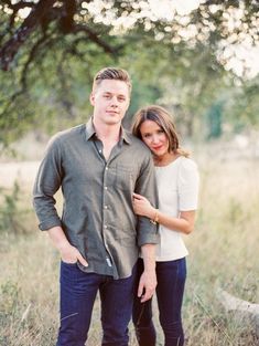 a man and woman standing next to each other in a field with trees behind them