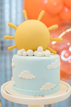 a blue and white cake topped with an orange sun on it's top, surrounded by balloons