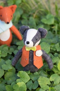 two small stuffed animals are sitting in the grass with green leaves around them and one is wearing an orange vest