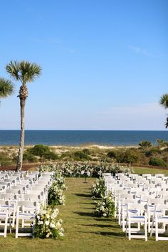 an outdoor wedding set up with white chairs and flowers on the grass by the ocean