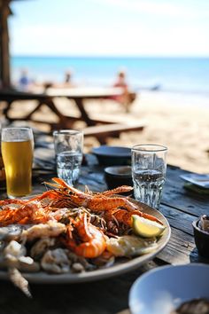 there is a plate of seafood and beer on the table at the beach with people in the background