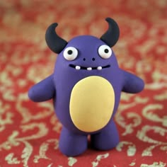 a purple toy with horns and eyes is on a red carpeted surface, holding an object in the shape of a ball