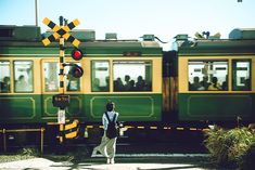 a person standing in front of a green and yellow train at a stop light with people walking by