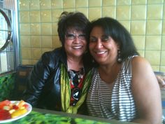 two women sitting next to each other in front of a plate of fruit on a table