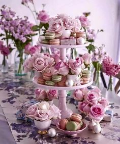 three tiered cake stand with pink roses and macaroons on it in front of purple flowers