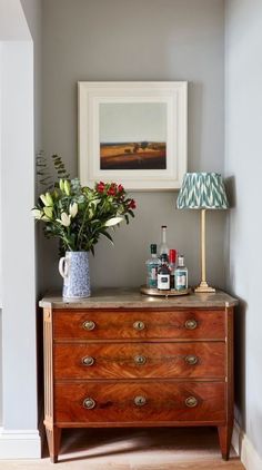 a dresser with flowers and bottles on it in the corner of a room next to a lamp