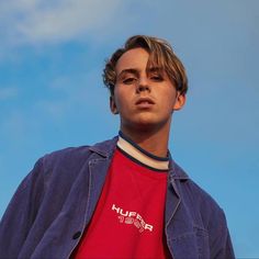 a young man wearing a red shirt and blue jacket with his eyes closed, standing in front of a blue sky