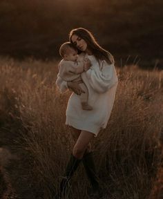 a woman holding a baby in her arms while standing in a field at sunset with the sun behind her