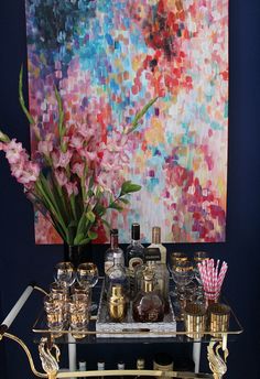 a bar cart with liquor bottles and glasses on it in front of a colorful painting