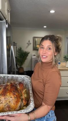 a woman holding up a turkey in a pan on the kitchen counter with an oven and refrigerator behind her