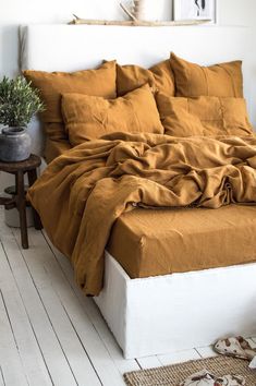 an unmade bed with brown sheets and pillows in a white room next to a potted plant