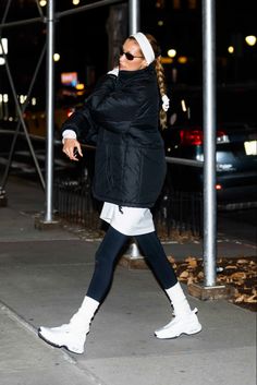 a woman in black jacket and white boots walking down the street at night with her hand on her hip