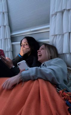 two women laying on a bed and one is holding a cell phone in her hand