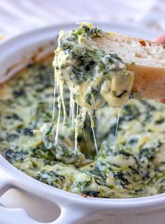 a hand holding a piece of bread over a bowl of spinach dip