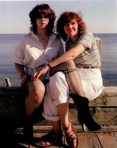 two women sitting on a wooden dock next to the ocean and one is holding her leg up