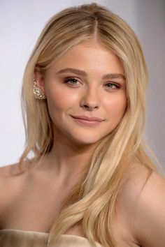 a woman with long blonde hair wearing a strapless dress and diamond earrings, smiling at the camera