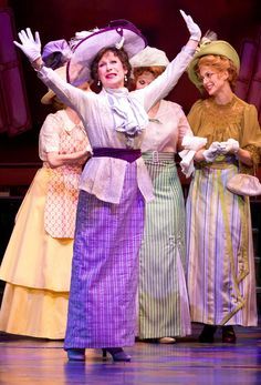 three women in dresses and hats on stage with their hands up to the side,