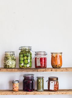 several jars filled with pickles and other food items on wooden shelves against a white wall