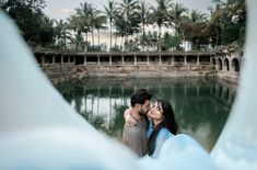 a man and woman are kissing in front of a lake with palm trees behind them