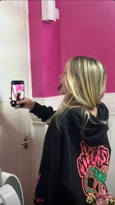 a woman taking a selfie with her cell phone in a pink bathroom stall while standing next to a urinal