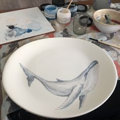 a white bowl with a whale painted on it next to paintbrushes and watercolors