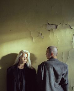 two people standing next to each other in front of a wall with peeling paint on it