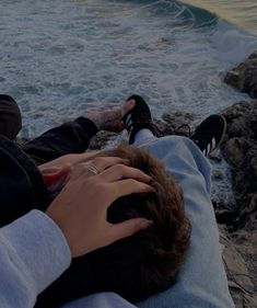 two people sitting on rocks near the ocean with their feet in each other's lap