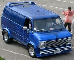 a blue van driving down a street next to a parking lot with two people in it