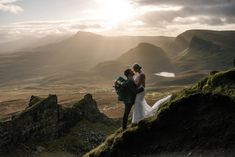 a bride and groom standing on top of a mountain with the sun shining through the clouds