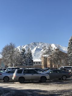 several cars parked in a parking lot with snow on the mountains behind them and buildings