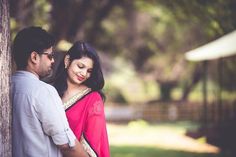 a man standing next to a woman in a red sari and white blouse, leaning against a tree