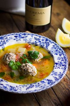 a bowl of soup with meatballs and vegetables next to a bottle of wine on a wooden table