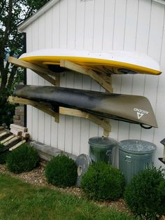 two surfboards mounted to the side of a white building next to bushes and trash cans