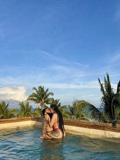 two women are sitting in the middle of a swimming pool with palm trees behind them