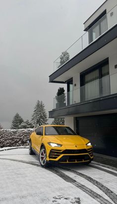 a yellow sports car parked in front of a house on a snow covered driveway with trees and bushes