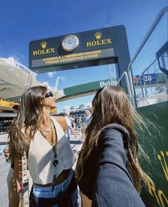 two women standing in front of a rolex sign at an outdoor event on a sunny day