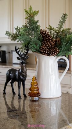 a white pitcher filled with pine cones on top of a counter next to a deer figurine