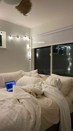 a laptop computer sitting on top of a bed covered in white sheets and blankets with lights strung across the windows