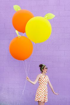 a woman holding three balloons in front of a purple wall with orange and yellow colors