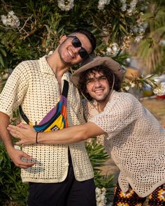 two men standing next to each other in front of some bushes and trees with flowers