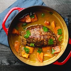 a pot filled with meat and carrots on top of a wooden table next to a blue napkin