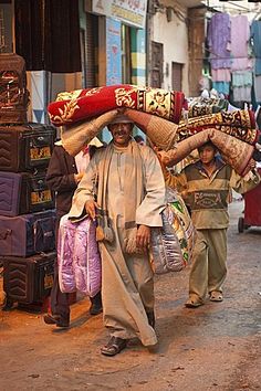 two men walking down the street carrying bags on their heads and suitcases on their shoulders