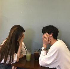 a man and woman sitting at a table with drinks in front of them, both holding their hands to their faces