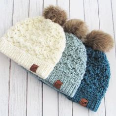 three crocheted beanies with pom - poms on white wood background