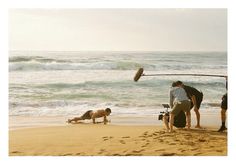 three people on the beach with one person falling off his surfboard and another man standing in the sand