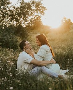 a man and woman are sitting in the grass with their arms around each other as the sun sets behind them