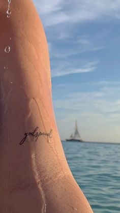 a person with a small tattoo on their arm near the water and a sailboat in the distance